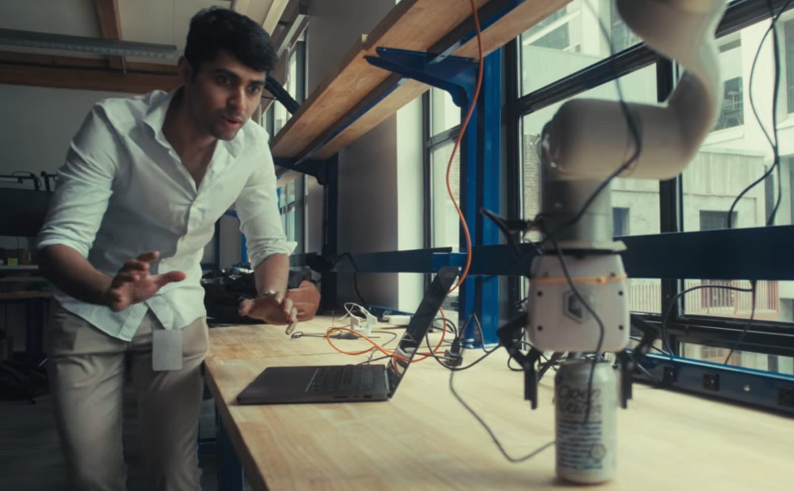 Muž v laboratoři sleduje robotické rameno, které na dřevěném pracovním stole uchopilo plechovku. Vedle leží notebook a spleť kabelů, v pozadí prosklená stěna dílny.